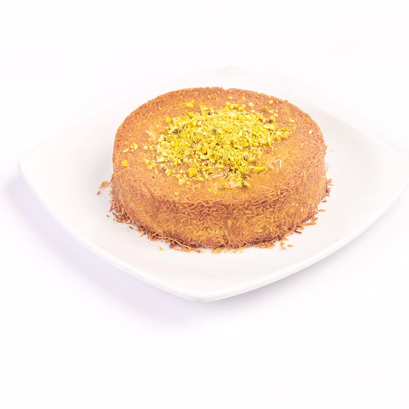Round kunafa dessert topped with crushed pistachios, served on a white square plate.