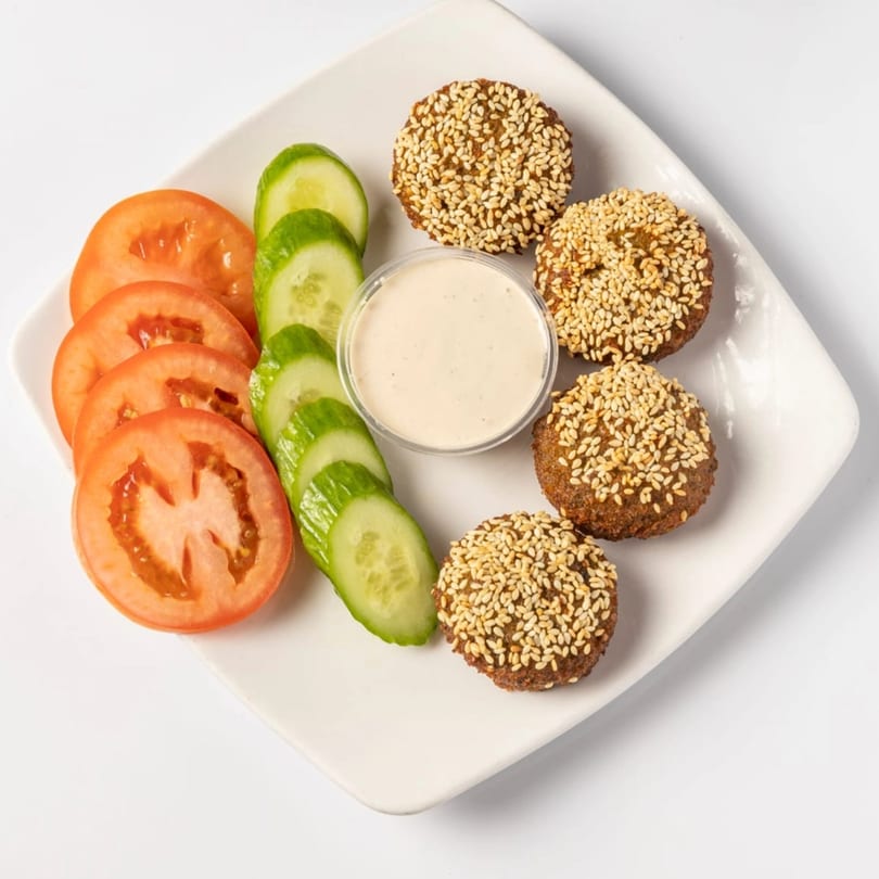 Kenz Cairo falafel with sesame seeds, sliced tomatoes, cucumbers, and a dipping sauce on a white plate.