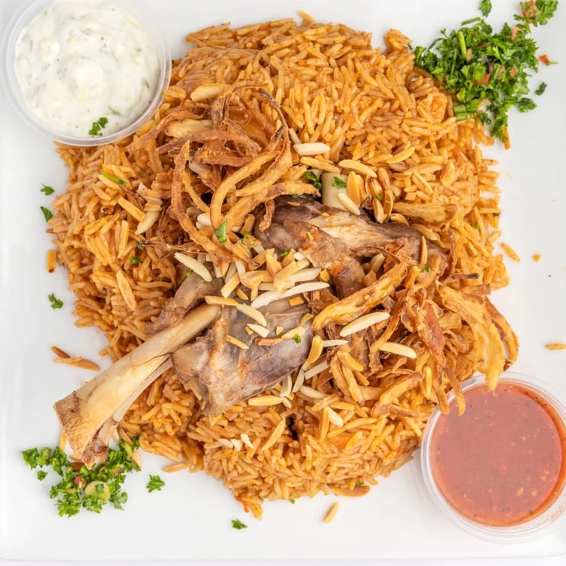 Middle-Eastern lamb shank with rice, fried onions, almonds, and two dipping sauces on a white plate.