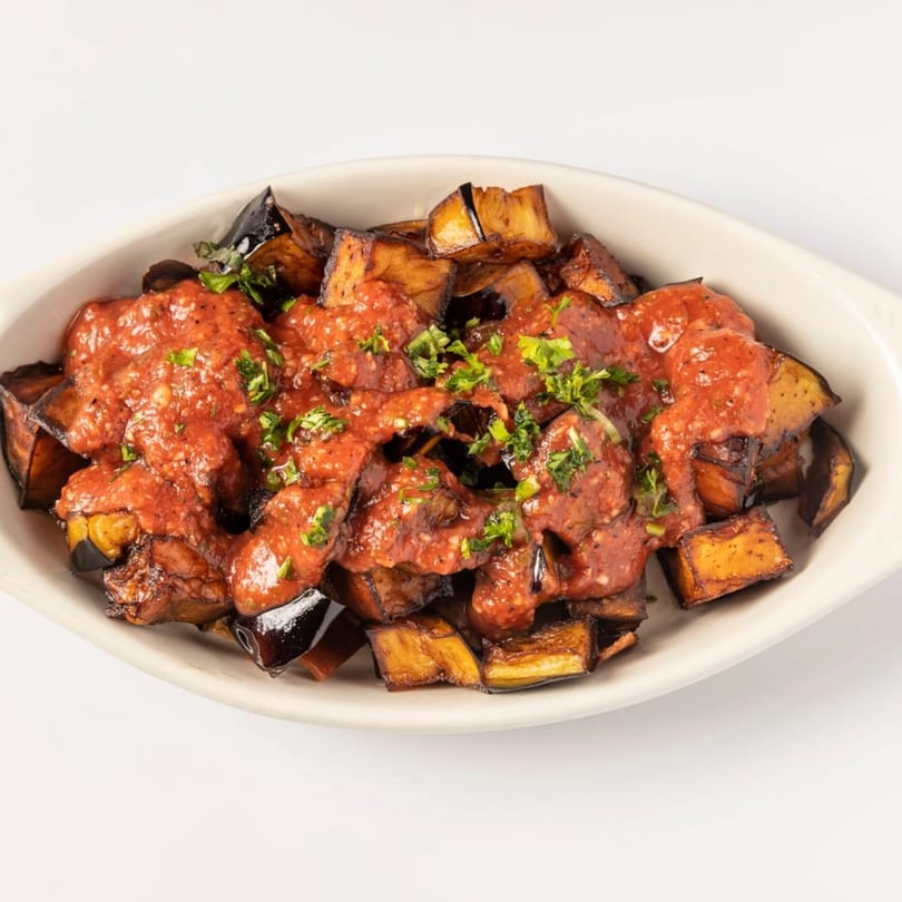 A white oval dish filled with roasted eggplant cubes topped with a rich red sauce and fresh parsley.