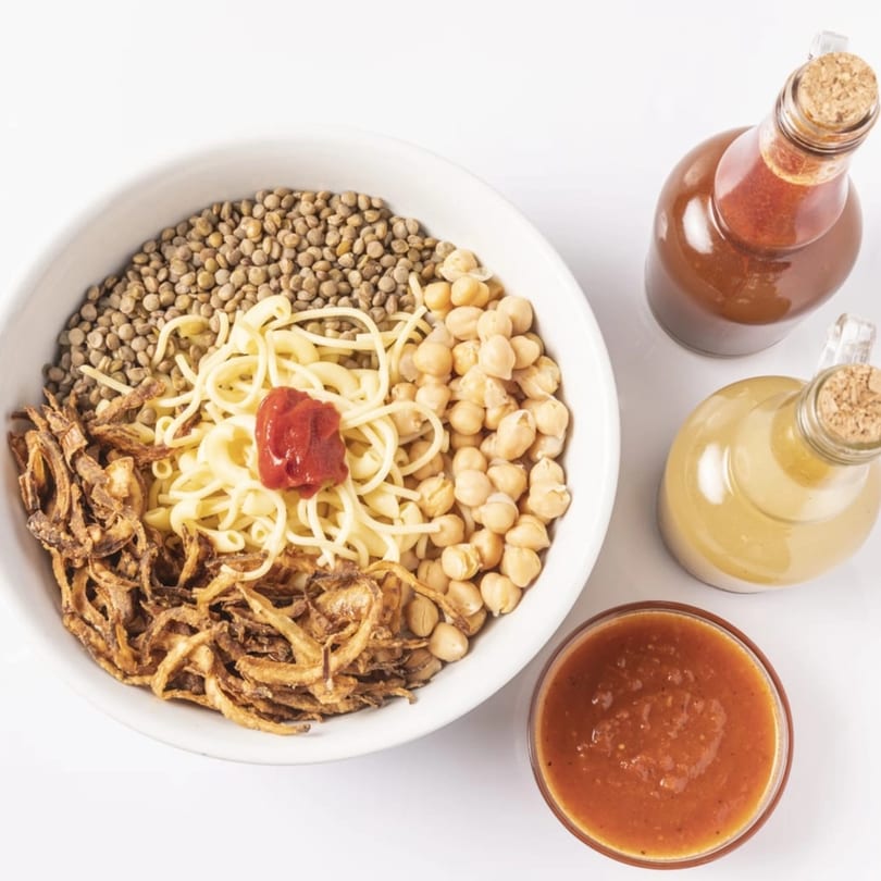 Koshary bowl with lentils, chickpeas, pasta, fried onions, and tomato sauce, with two sauce bottles.