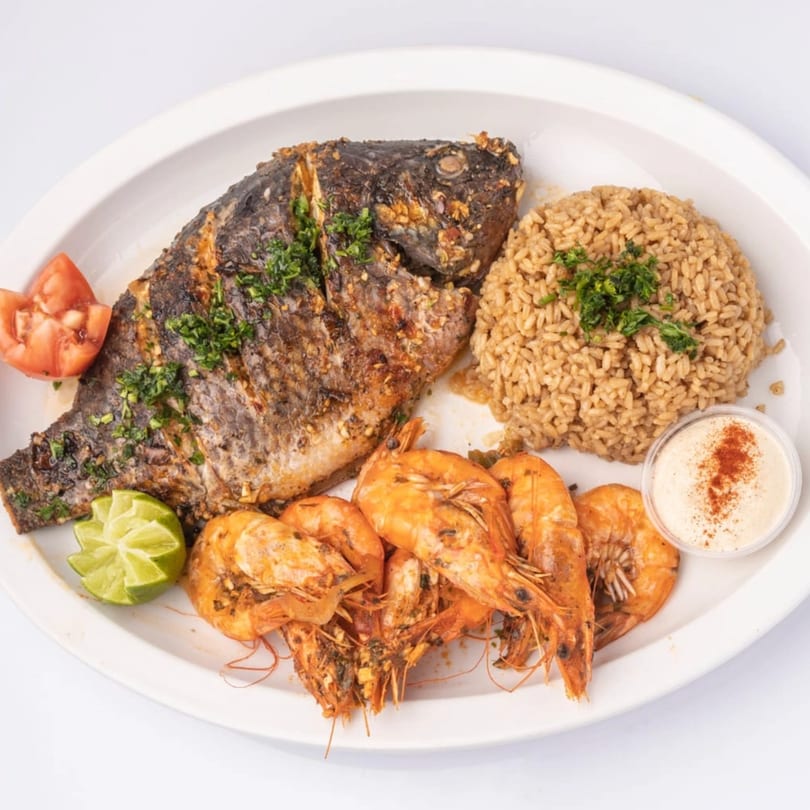 Grilled whole fish, shrimp, rice, lime, tomato, and a dipping sauce on a white plate.